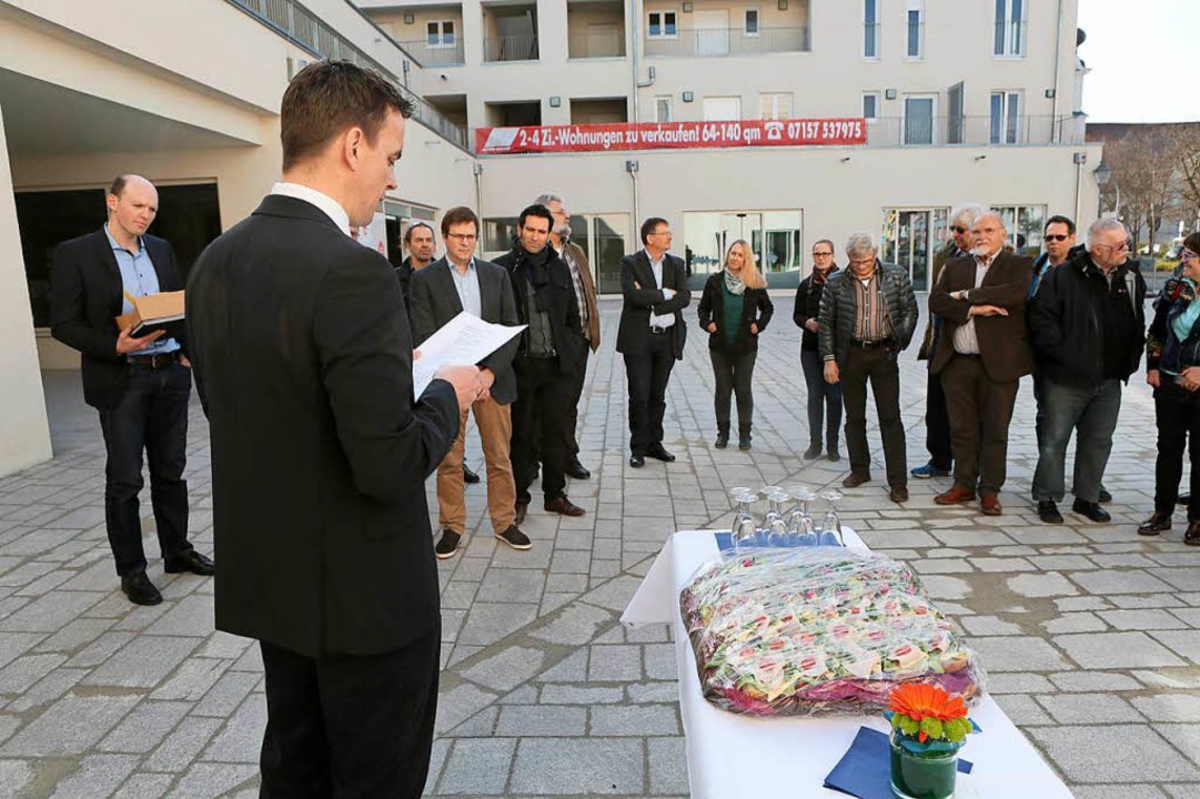Mann in schwarzem Anzug liest vor einer Gruppe von etwa 15 Personen im Innenhof eines Wohngebäudes mit Banner für Wohnungsverkauf, auf Tisch im Vordergrund Blumenstrauß und mehrere leere Weingläser