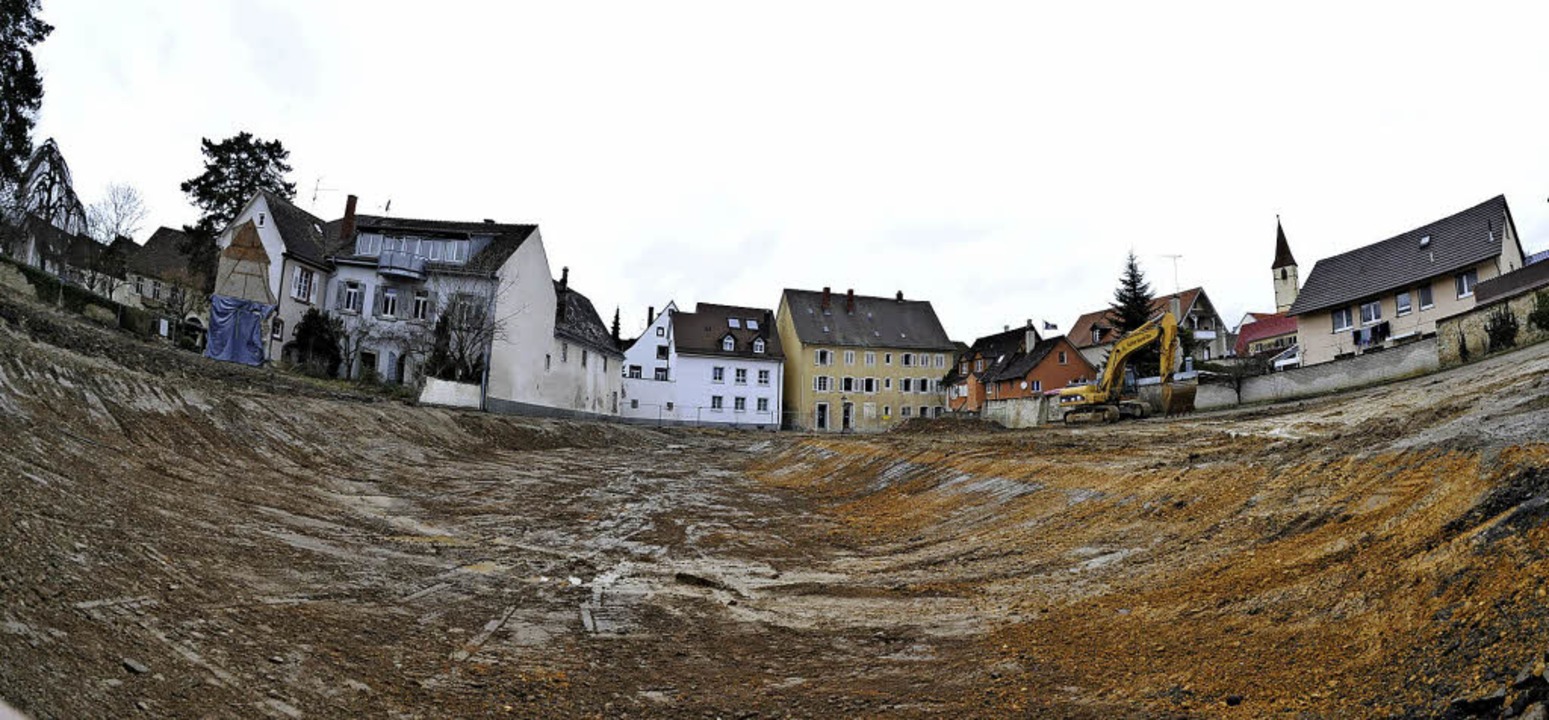 Große Baugrube in einem Wohngebiet mit Abriss von Gebäuden im Hintergrund und Bagger auf der Baustelle unter bewölktem Himmel