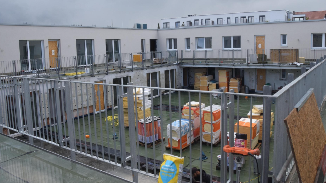 Innenhof einer modernen Wohnanlage in Bauphase mit flachem Dach, Stapeln von Isolationsmaterialien und Baumaterialien, umgeben von weißen Gebäuden mit großen Fenstern und Baustellenabsperrungen